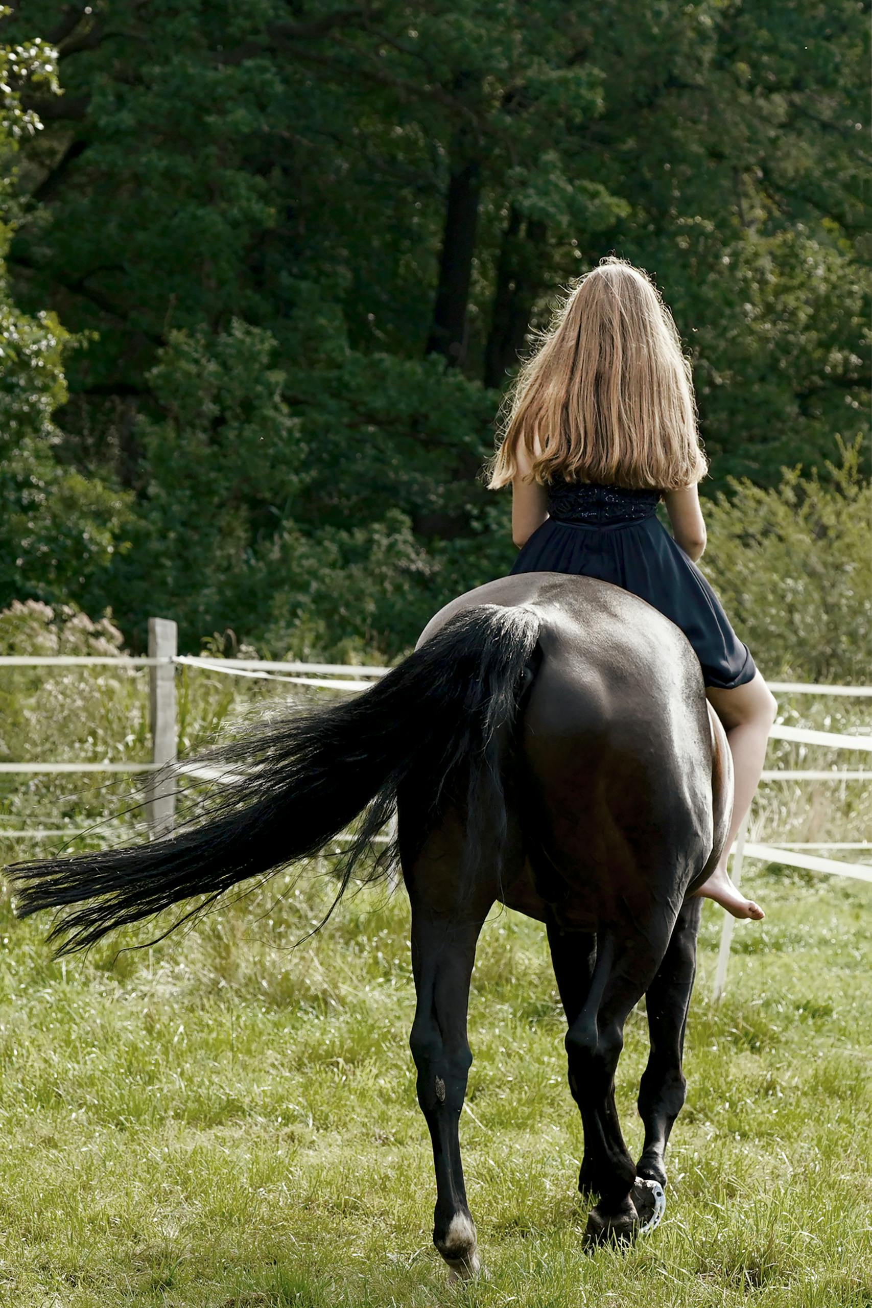 A woman with blonde hair riding a black horse in a green field with a fence.